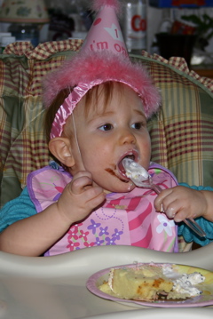 Domino, sitting in her high chair, wearing a party hat, eating cake and ice cream.